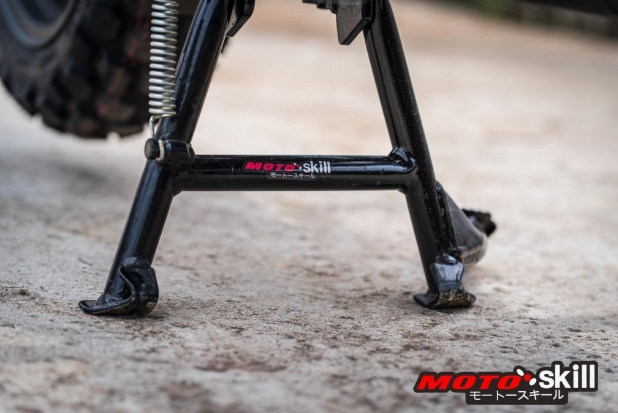 Close-up of a motorcycle kickstand with 'Moto Skill' branding on a concrete surface.