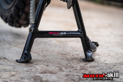 Close-up of a motorcycle kickstand with 'Moto Skill' branding on a concrete surface.
