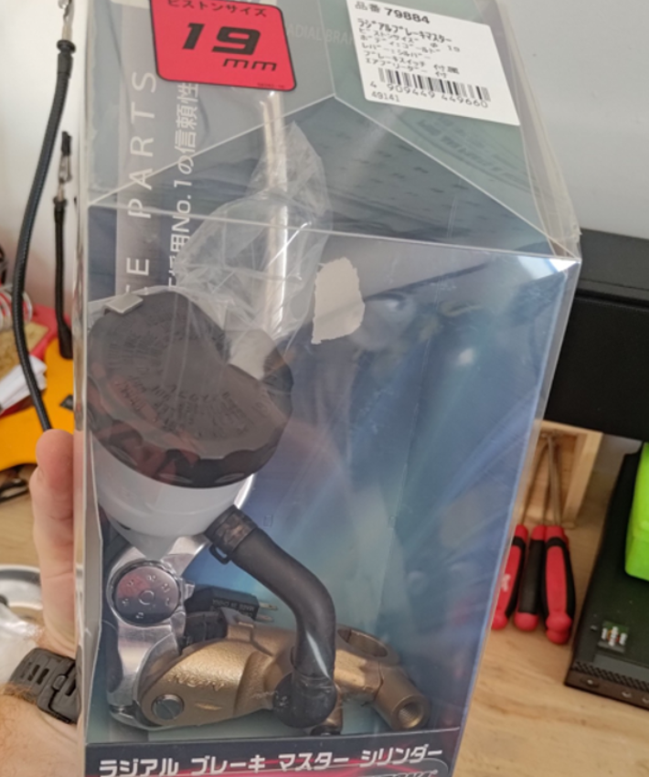 Product packaging with a clear window showing a mechanical part, held by a hand.